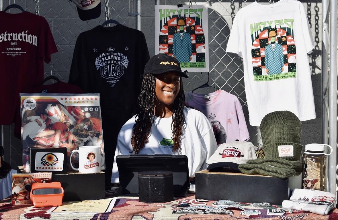 Woman sitting behind a table at Hazy Fest event, wearing a black cap, with merchandise including t-shirts, hats, and a green knit hat on display, background shows a metal fence and event poster.