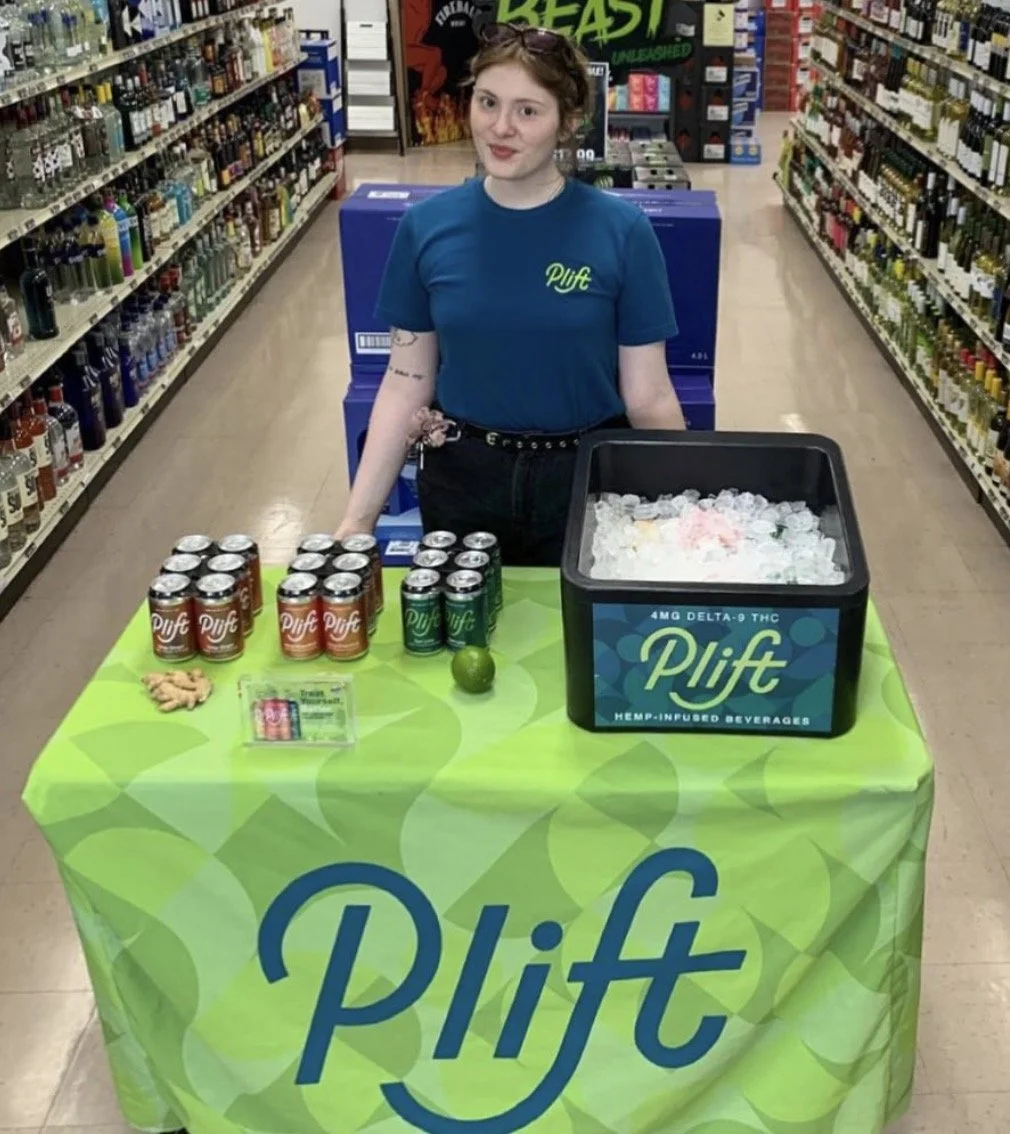 A young woman standing behind a table with a green tablecloth displaying the brand name Plift. On the table, there are canned beverages, a lime, a small pile of ginger, and a container filled with ice and Plift drinks. She is in a store aisle, wearing a blue t-shirt with the Plift logo, with shelves stocked with various bottles in the background.