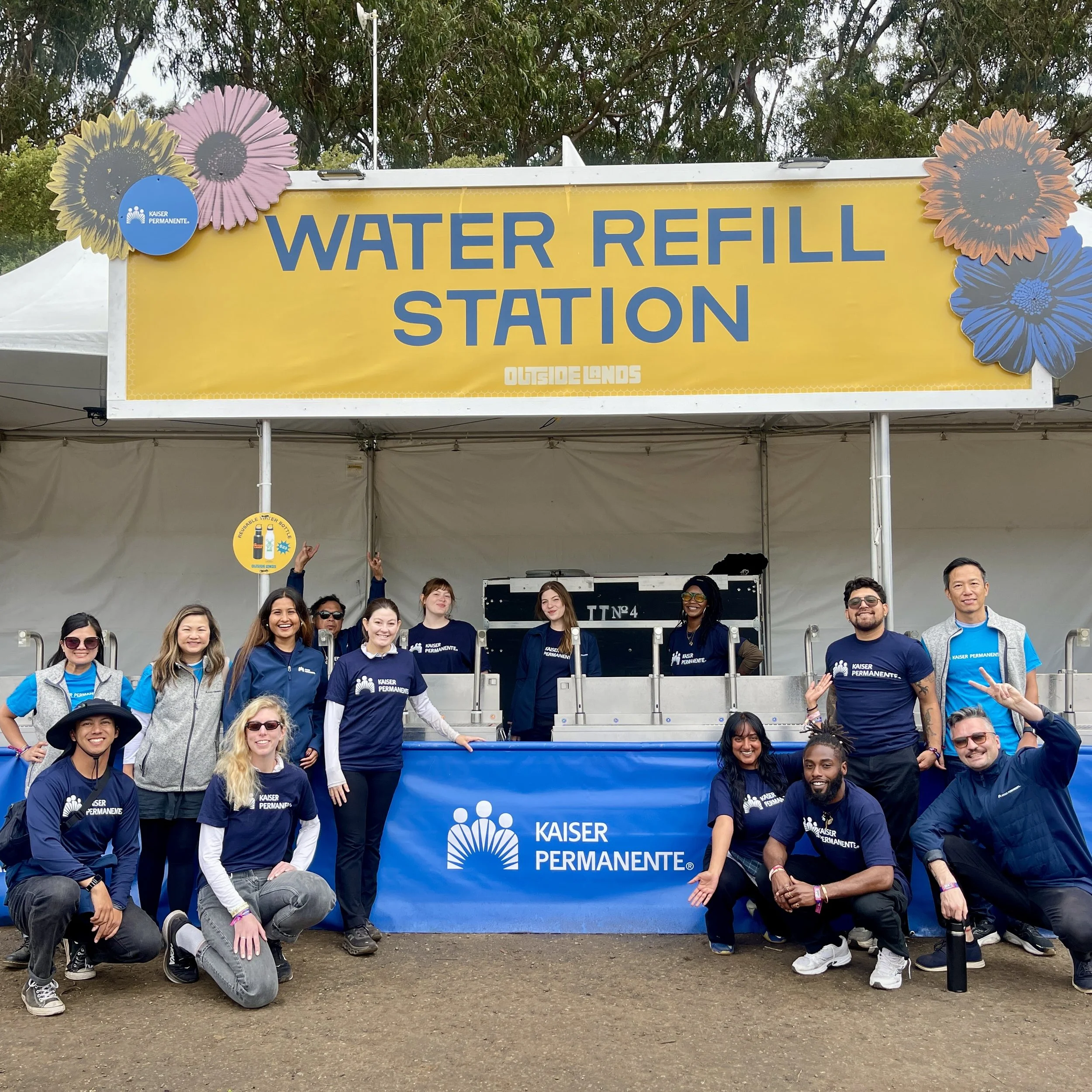 Group of smiling people standing in front of a yellow sign that reads Water Refill Station with the Kaiser Permanente logo, surrounded by paper flower decorations, outdoors with trees in the background.