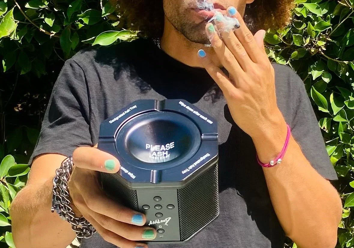 Person smoking vapor from a vaporizer, holding a portable speaker, standing amidst green leaves.