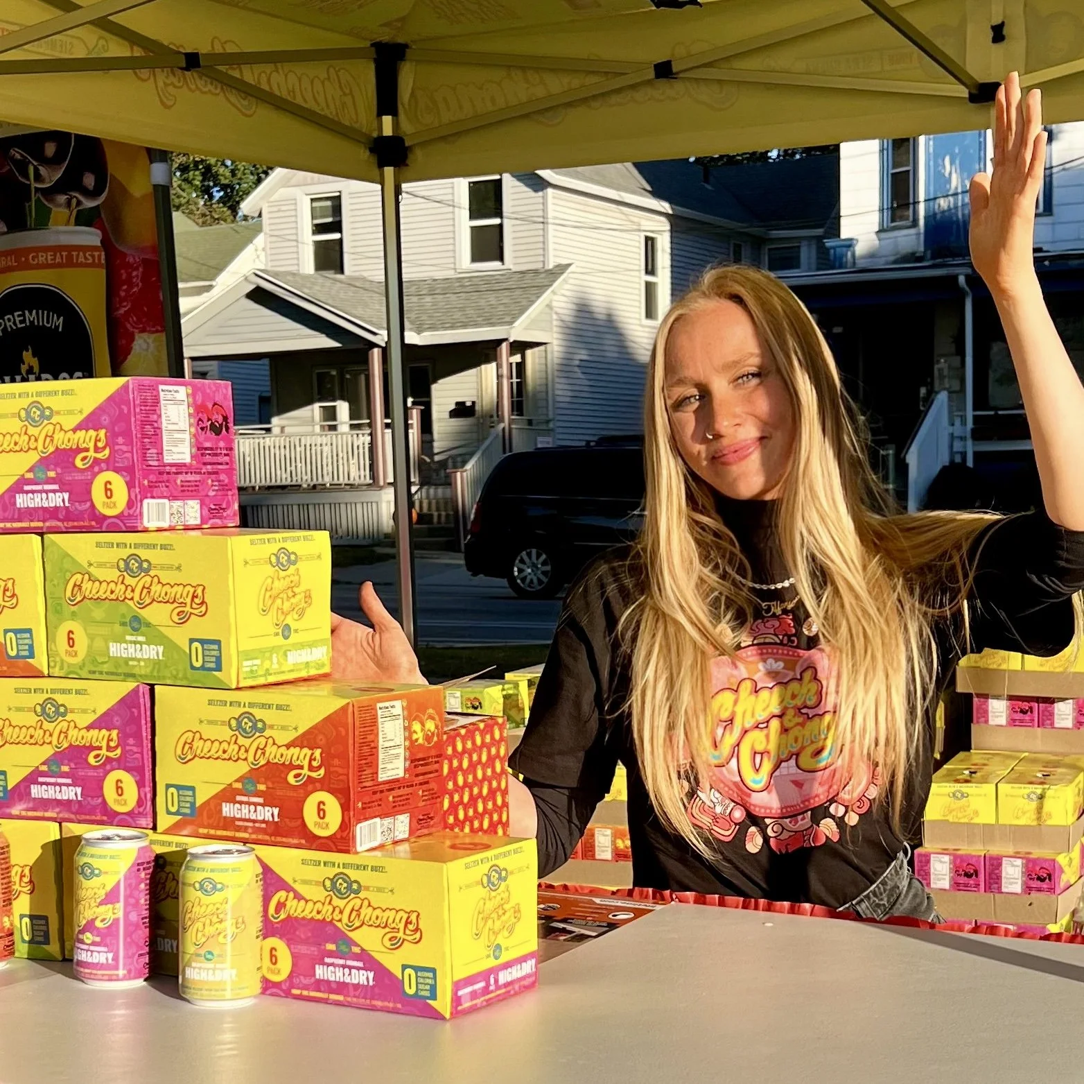 A woman with long blonde hair smiling at a snack stand under a yellow canopy, with boxes of Chuckles Chums in front of her and houses in the background.