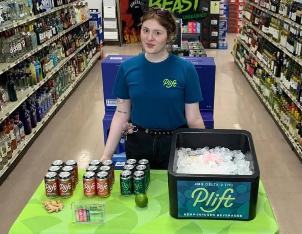 A young woman in a blue shirt with 'Plift' written on it, standing behind a table with canned beverages and a bucket labeled 'Plift' containing ice and cannabis products, in a grocery store aisle.