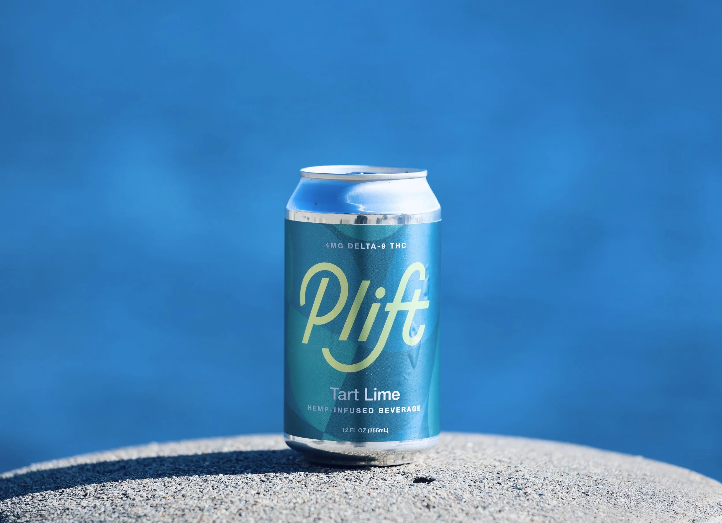 A tall light blue can of Delta-9 THC beverage with the flavor 'Tart Lime' written on it, placed on a textured surface outdoors with a blue sky in the background.