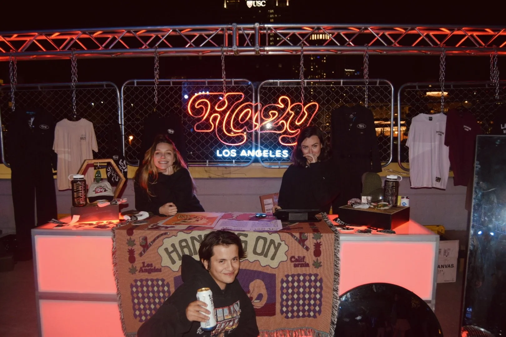 Nighttime scene of a food truck in Los Angeles featuring two women behind the counter and a young man holding a can in front. The truck is decorated with a Los Angeles sign, hanging T-shirts, and a colorful blanket on the counter. Neon lights illuminate the scene.