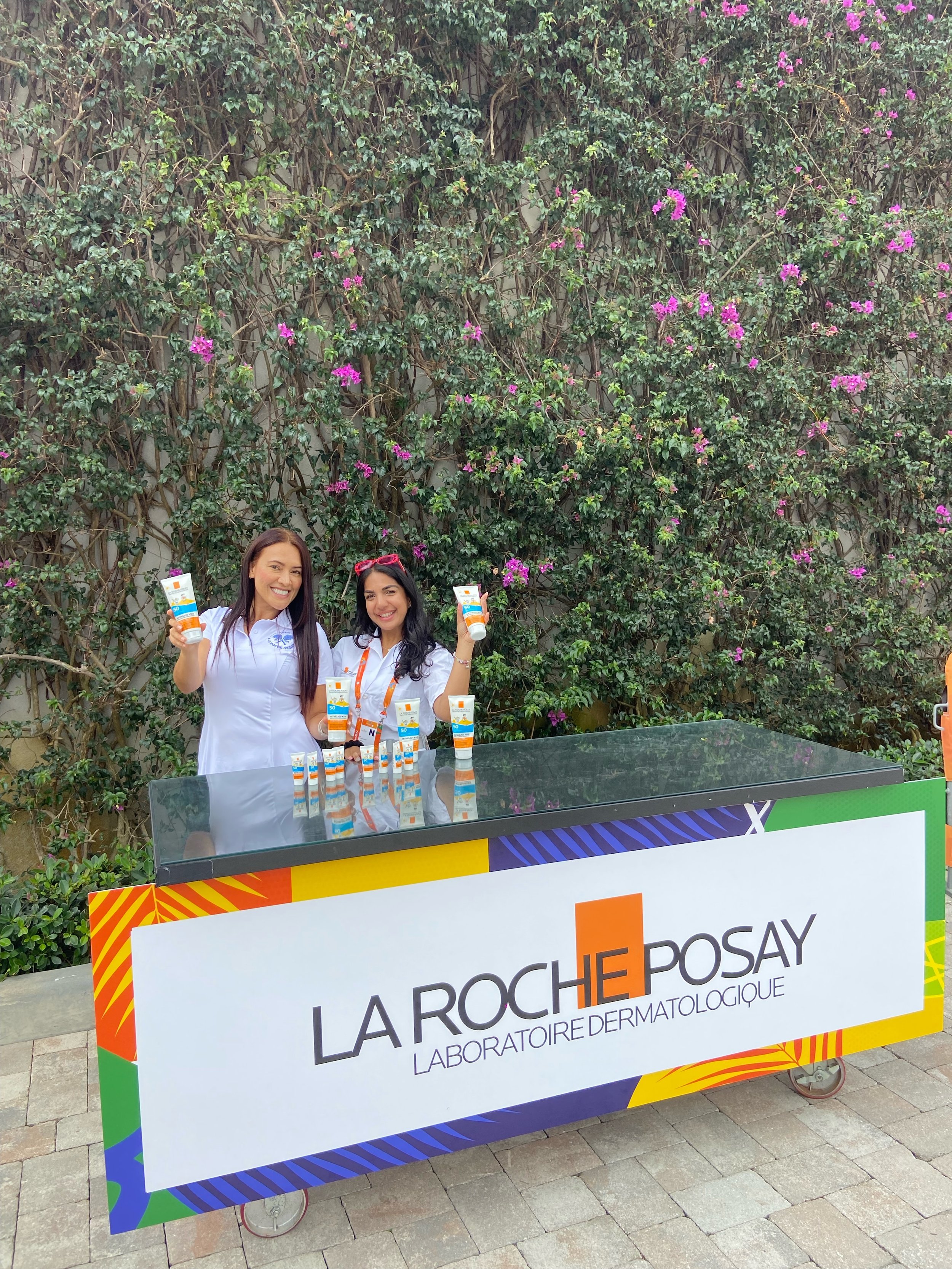Two women standing behind a table with skincare products, smiling and holding products at an outdoor promotional event for La Roche-Posay, with a colorful sign and a leafy green wall with pink flowers in the background.