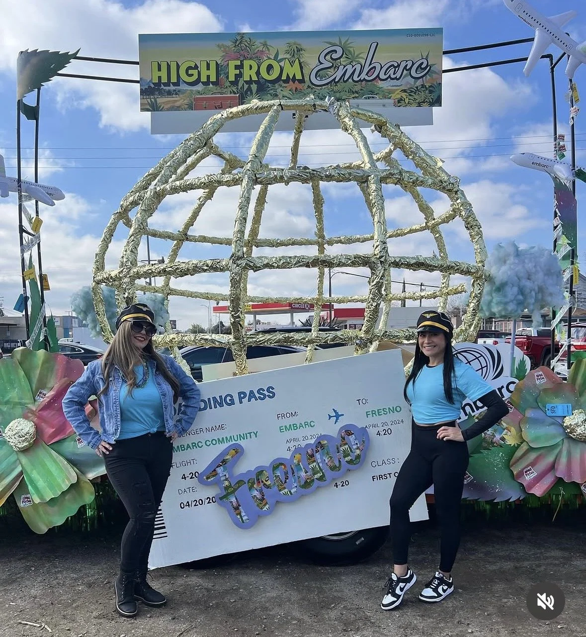 Two women in flight attendant costumes standing in front of a float decorated like a globe, with a banner that has a boarding pass design and the word 'Thankful' on it. The float is part of a parade or celebration.