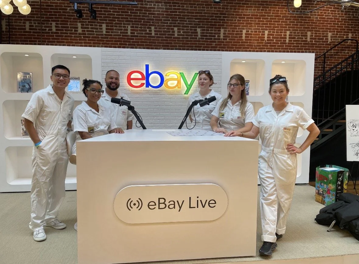 A group of six people standing behind a white desk with microphones, in front of a white wall with the eBay logo in colorful letters, in an indoor setting resembling a studio or office.