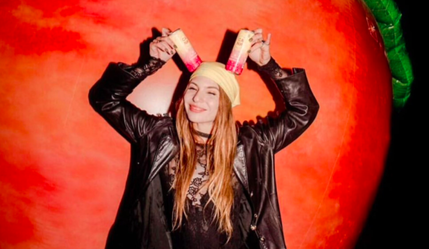 A woman with long hair, wearing a black lace top, black leather jacket, and yellow headscarf, smiles as she holds two cans of soda on her head with a large red and green inflatable background.