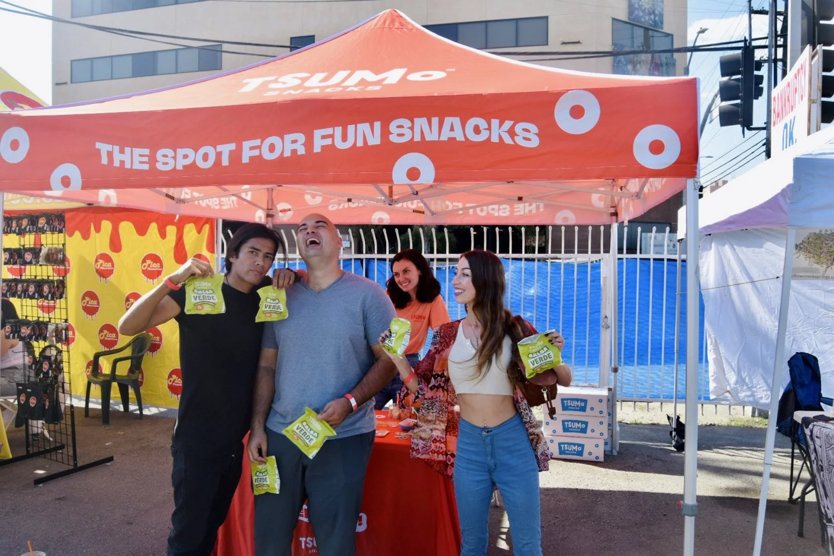 People holding snack bags at an outdoor booth with a red canopy titled 'The Spot for Fun Snacks.'