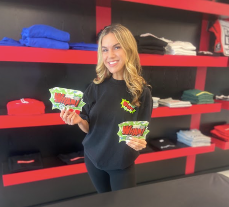 A young woman smiling and holding two comic book style signs that say 'WHAM!' in a retail store with shelves of folded clothes in the background.