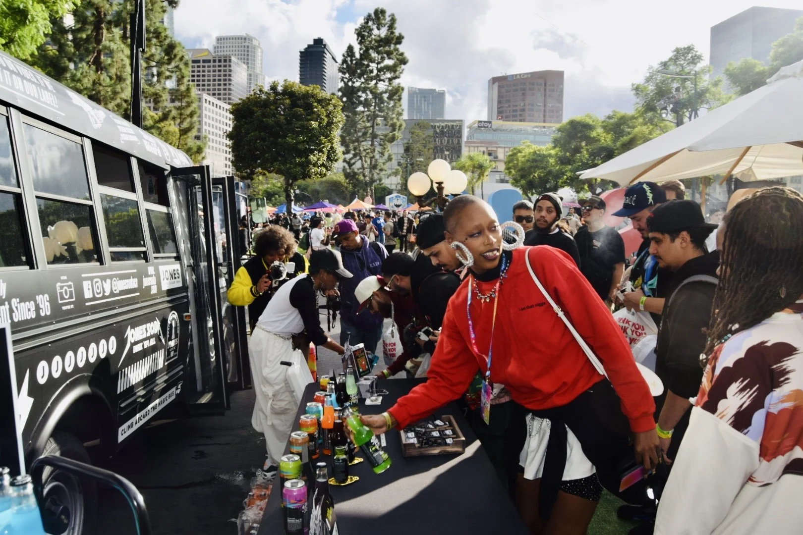 A diverse group of people gathered at an outdoor event in a park with city buildings in the background. The scene includes a woman in a red jacket browsing bottles of beverages on a table next to a black bus, with others mingling under umbrellas and tents on a sunny day.