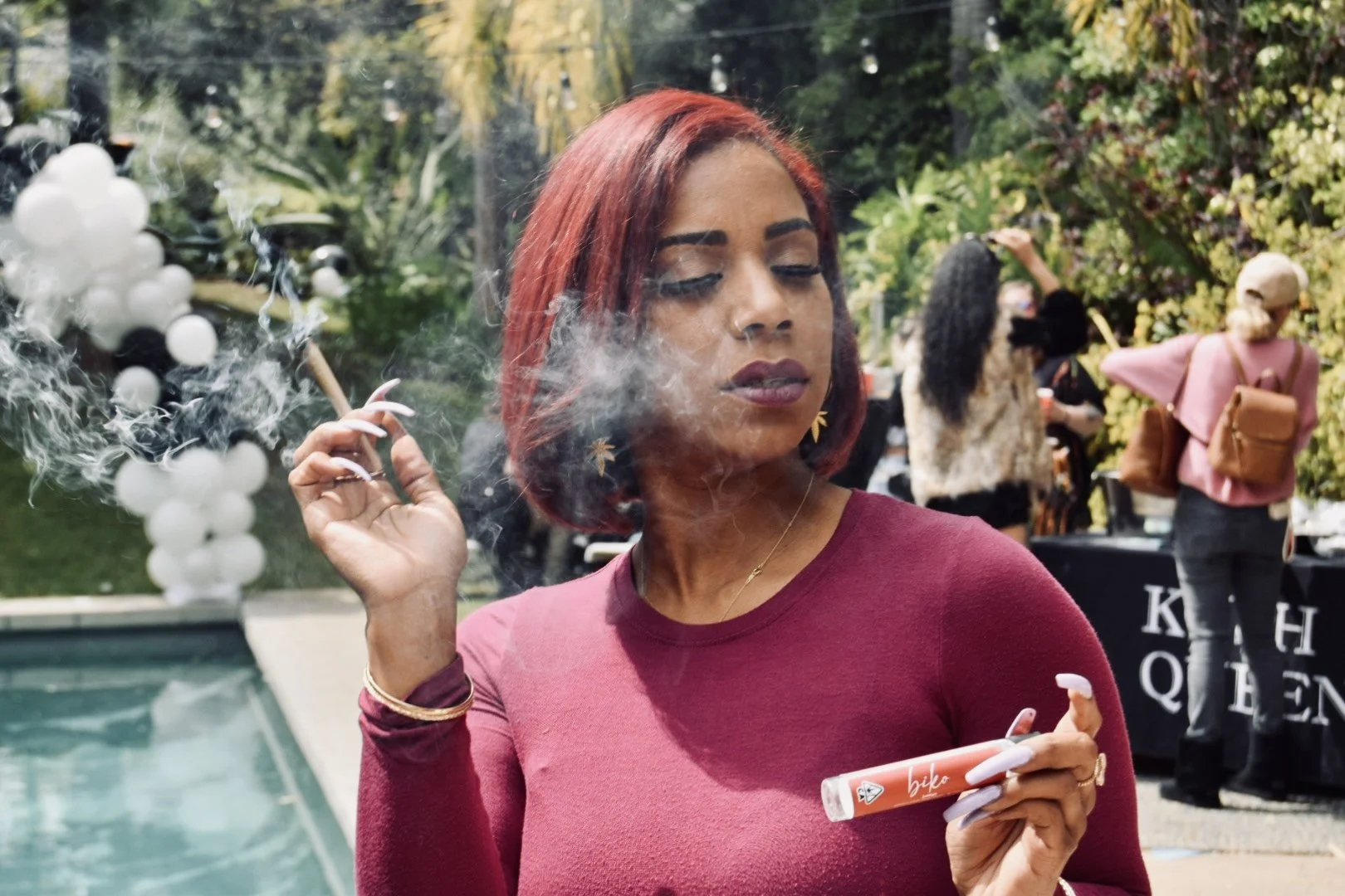 Woman with red hair holding a vape and an orange lip gloss tube outdoors with other people in the background.