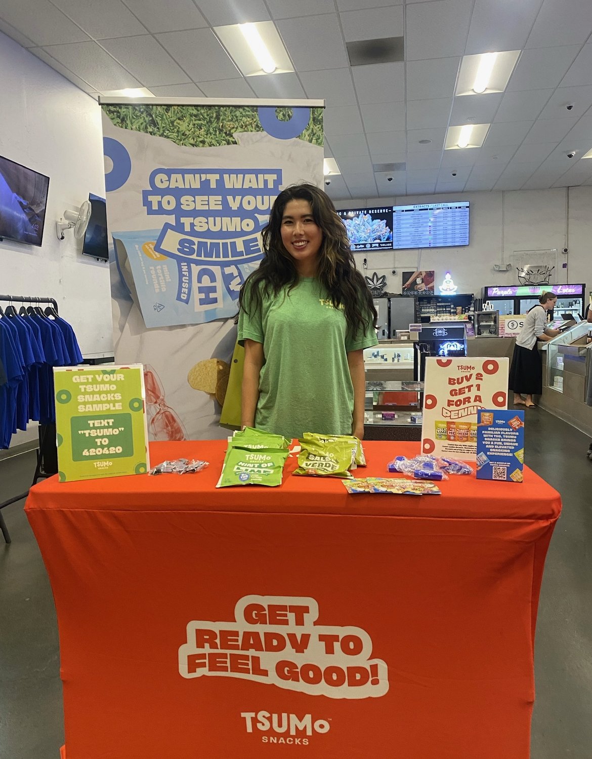 A woman standing behind an orange table at a TSUMo snacks booth inside a retail store. The table displays green and yellow snack packages, promotional signs, and a QR code. She is smiling and wearing a green TSUMo t-shirt.