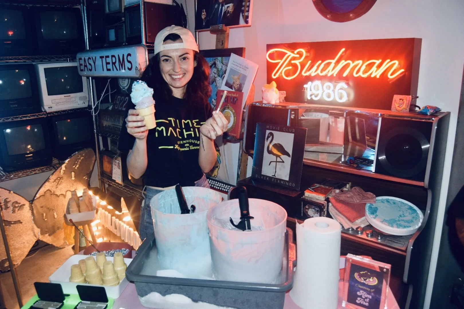 A woman is smiling while holding an ice cream cone and a spoon, standing in front of a colorful, retro-themed setup with neon signs, vintage television screens, and various items on display, including a poster of a black flamingo and a box labeled "Movie Club."