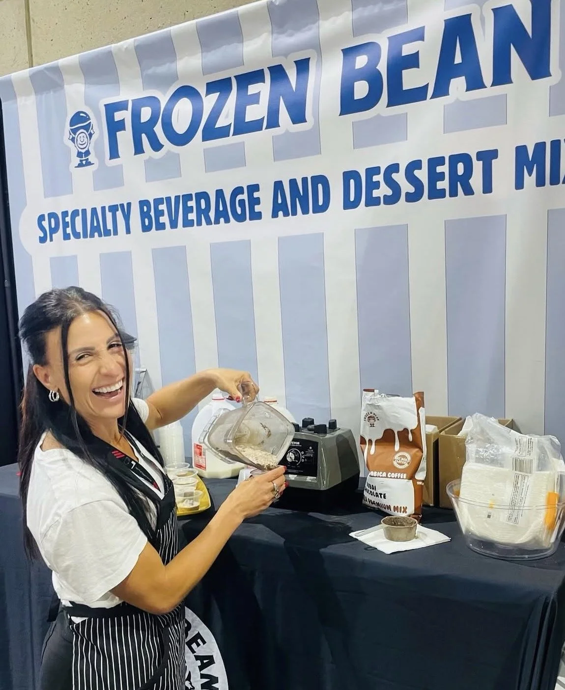 A woman in a black and white striped apron smiling and pouring frozen beans into a container at a booth with large sign 'Frozen Bean, Specialty Beverage and Dessert Mix'.