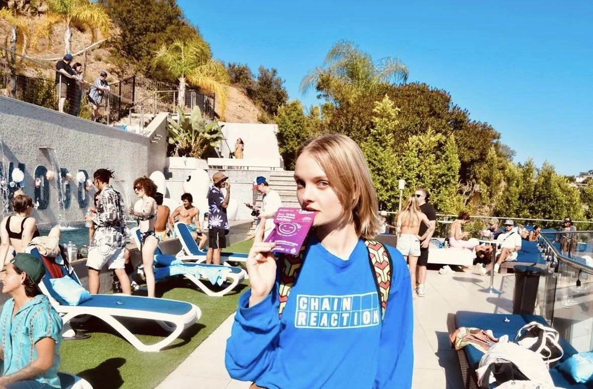 Young woman with shoulder-length blonde hair holding a purple packet near her face at a busy outdoor pool area, surrounded by lounge chairs, people sunbathing and socializing, with water features, lush greenery, and clear blue sky in the background.
