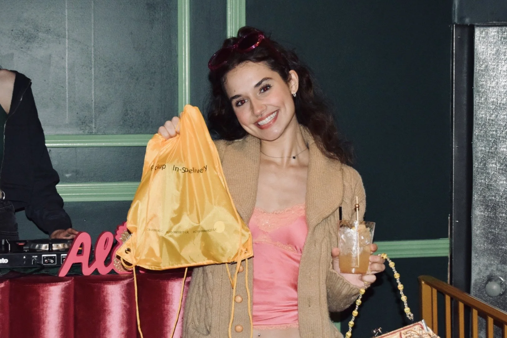 Young woman smiling and holding a drink and a yellow shopping bag indoors with dark green walls.