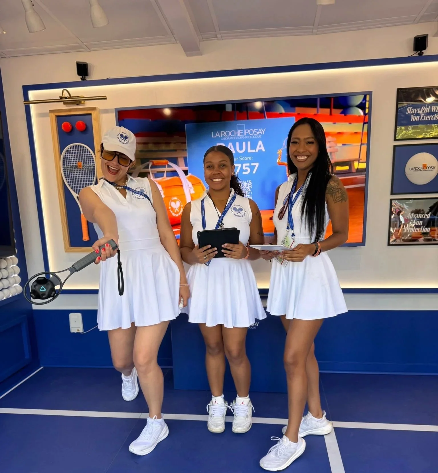 Three women in white sports dresses standing on a blue court, smiling at the camera during an event, with a large La Roche-Posay sign in the background.
