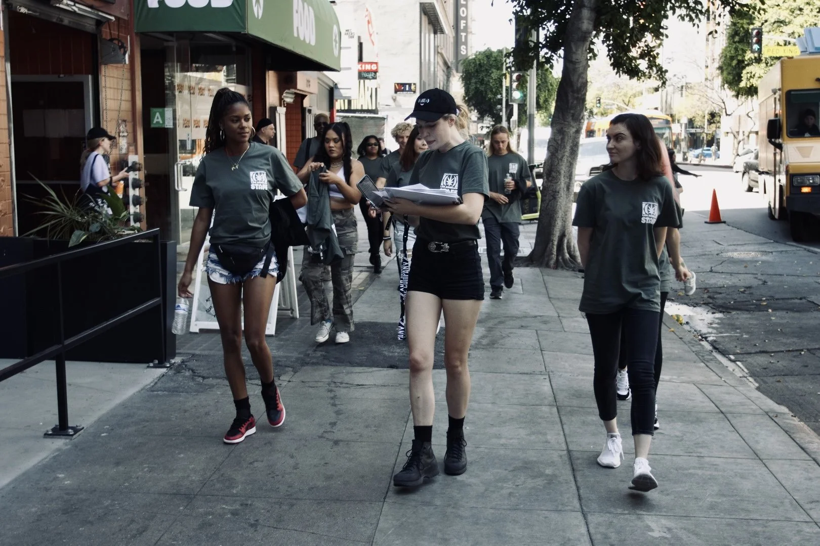 A group of young women walking on a city sidewalk, some holding water bottles, wearing matching gray T-shirts with a UN logo, suggesting they are part of a volunteer or organization group.