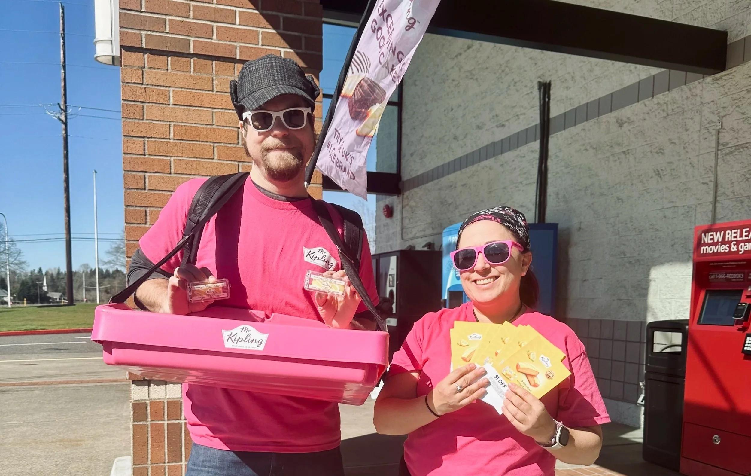 Two smiling people, a man and a woman, wearing pink shirts and sunglasses, standing outside a store near a red newspaper vending machine. The man is holding pink swag items, and the woman is holding yellow flyers. There is a brick wall, a sign, and a blue sky in the background.