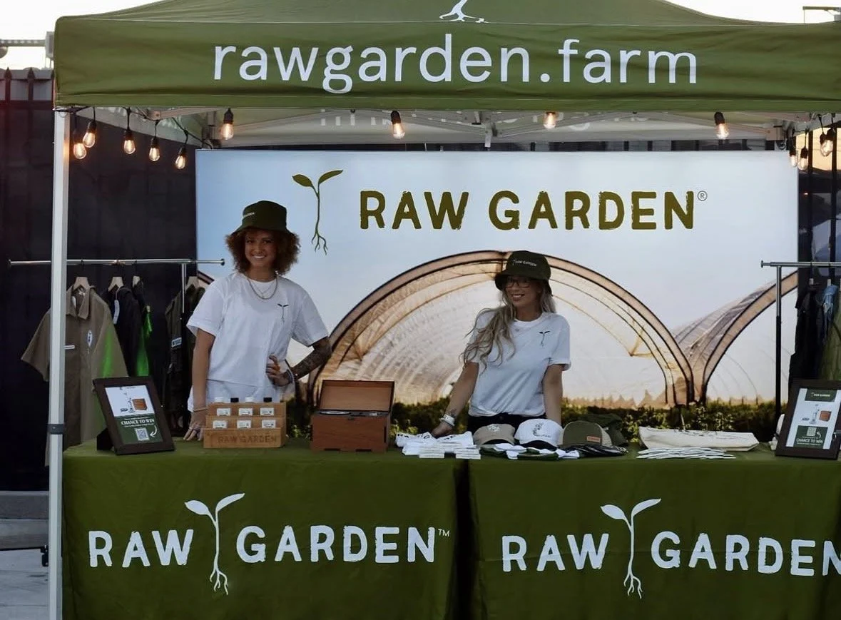 Two women standing behind a booth with signs for RAW GARDEN and rawgarden.farm, selling merchandise and products at an outdoor event.