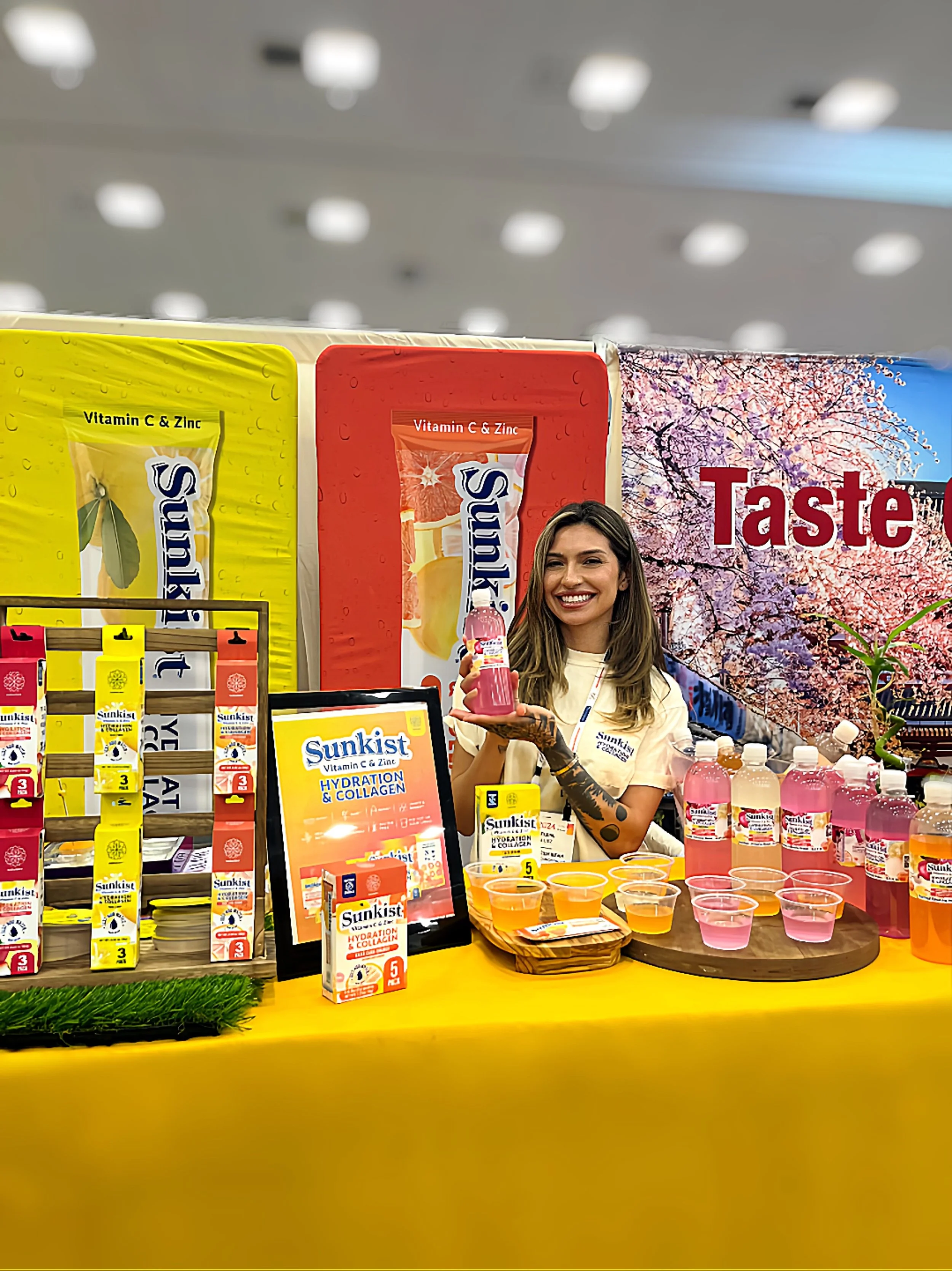 A woman smiling at a booth displaying Sunkist vitamin C and zinc hydration and collagen drinks, with various flavors in bottles and small cups, and promotional signage in a large indoor setting.