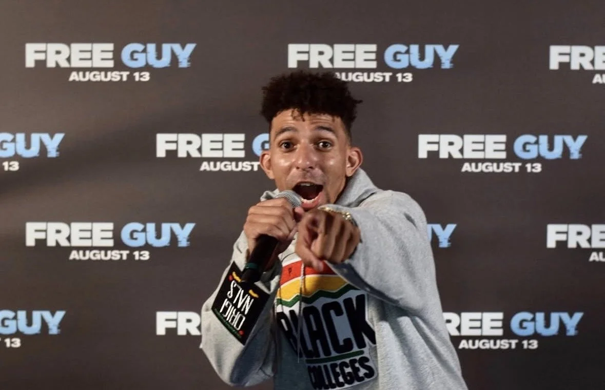 A man holding a microphone, pointing towards the camera and smiling, standing in front of a backdrop that reads 'Free Guy August 13' repeatedly.