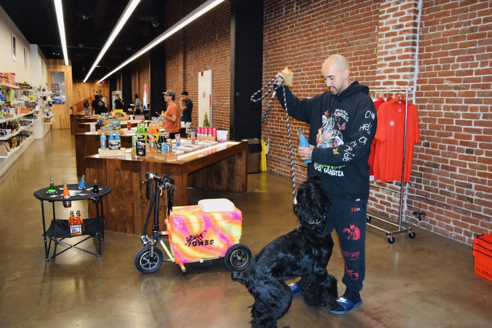 A man with a shaved head holding a leash attached to a large black dog in a retail store. The store has exposed brick walls, wooden counters, and shelves with various products. Other customers are browsing in the background.