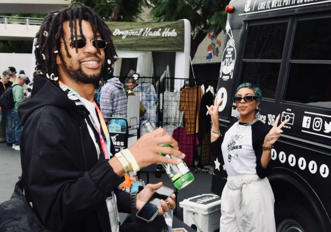 A man with dreadlocks wearing sunglasses, a black hoodie, and wristbands, holding a drink and a phone. A woman with short hair, sunglasses, and a peace sign gesture, standing next to a black food truck with white signage. People are in the background at an outdoor event with vendor tents.
