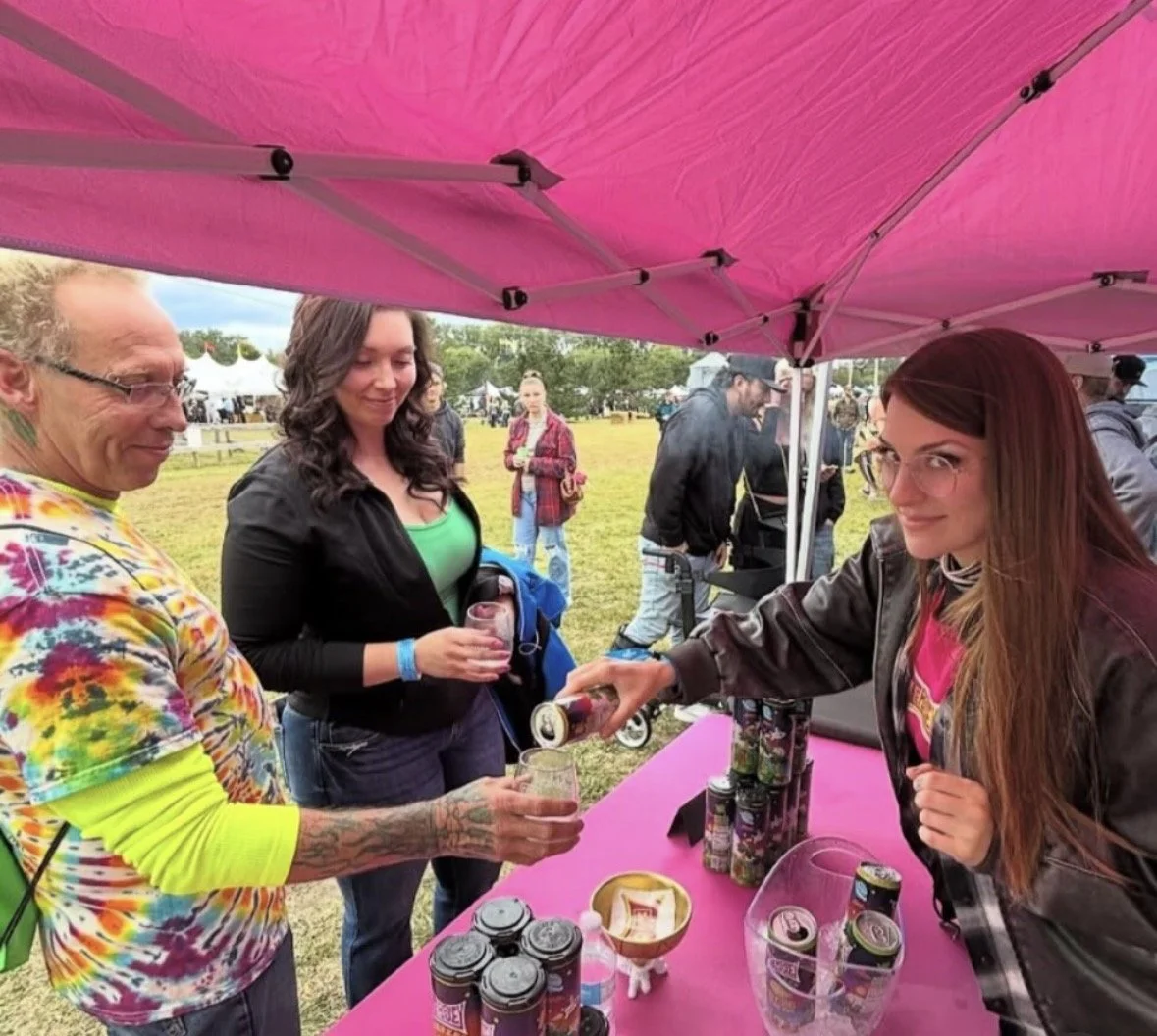 People at a pink canopy tent selling canned beverages at an outdoor event, with one person pouring a drink for another.