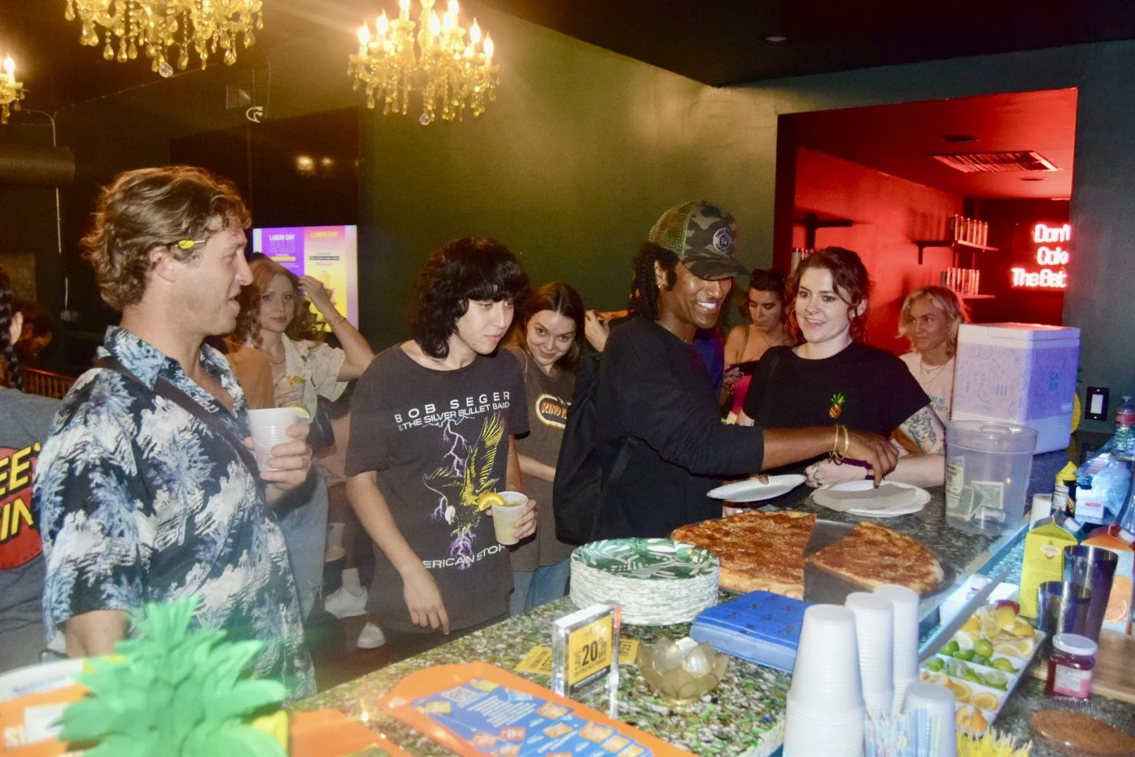 People in line at a pizza stand, some holding drinks, in a dimly lit room with chandeliers and neon signs.