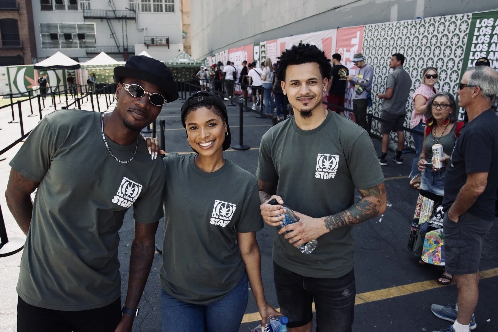 Three people standing together at an outdoor event, wearing matching gray T-shirts with a logo that says 'Los Angeles Staff.' They are smiling and posing for the photo, with several other people and event booths in the background.