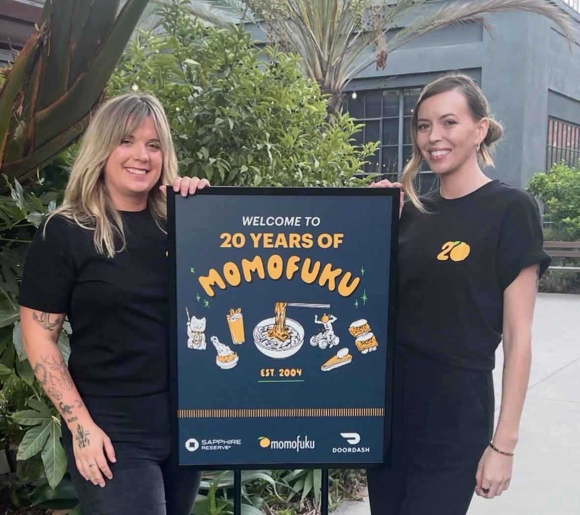 Two women standing outdoors, holding a sign celebrating 20 years of Momofuku, with greenery in the background.