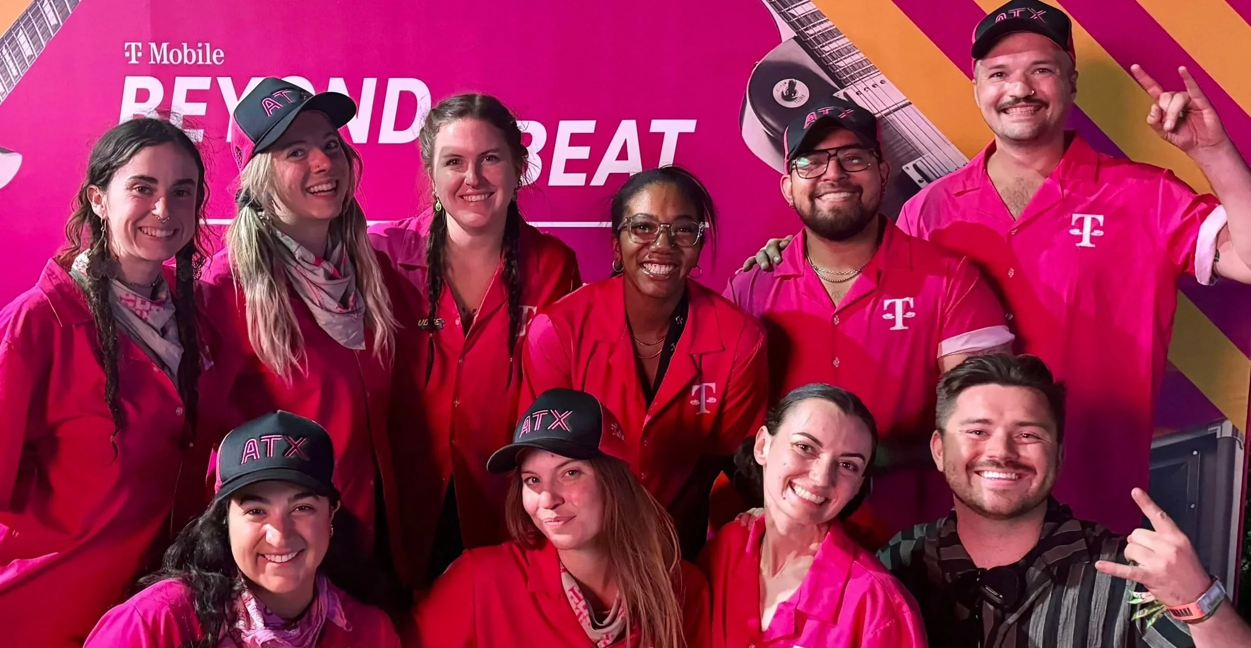 Group of diverse people smiling and taking a group photo at a T-Mobile event, with pink and orange background featuring guitars and the words 'Beyond' and 'BEAT.' They are all wearing pink uniforms, some with hats and bandanas.
