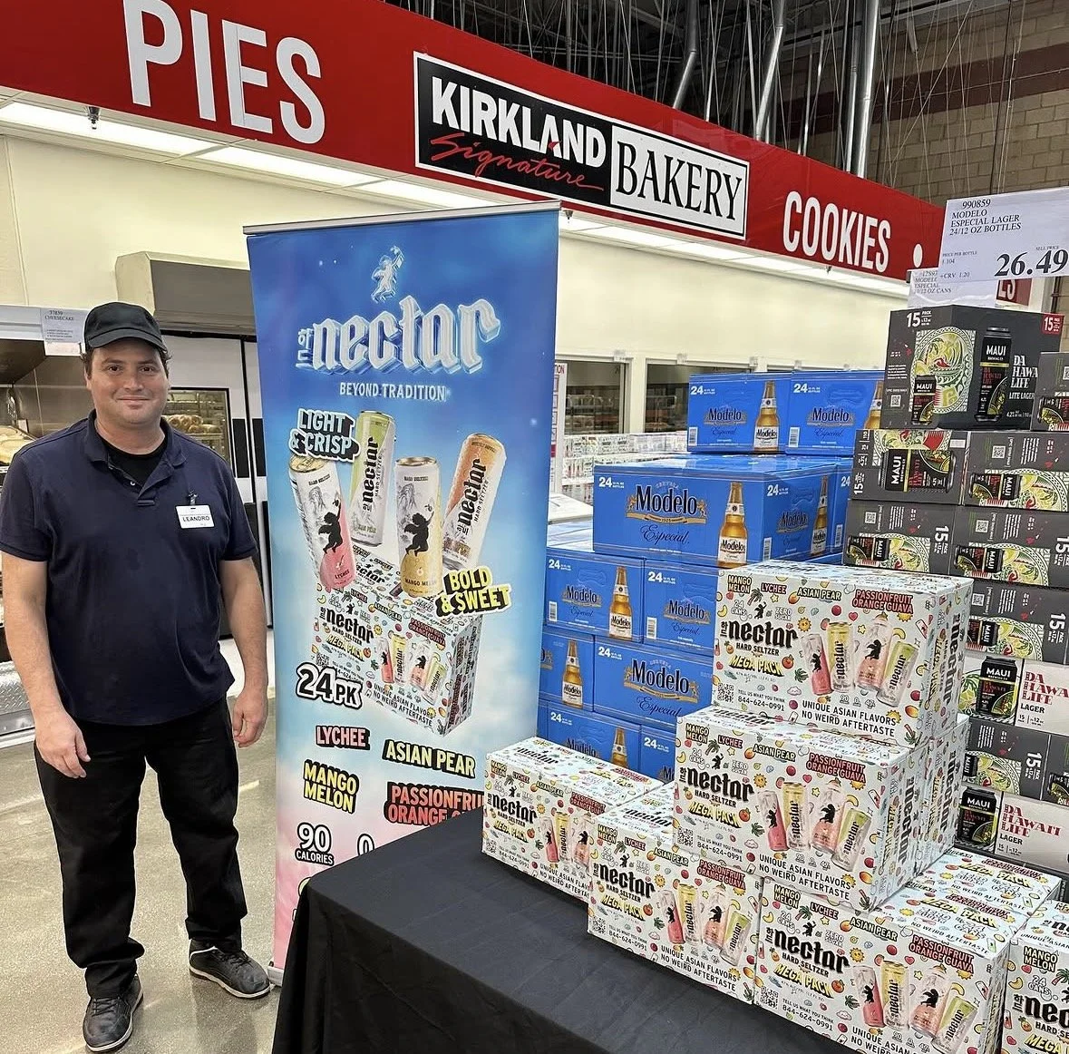 A man standing next to a display of Nectar hard seltzer drinks at a store. The display features multiple boxes of Nectar flavors, including lychee, Asian pear, mango melon, and passionfruit orange guava. Behind the man, there are stacked cases of Modelo beer, and signage indicating prices and product information.