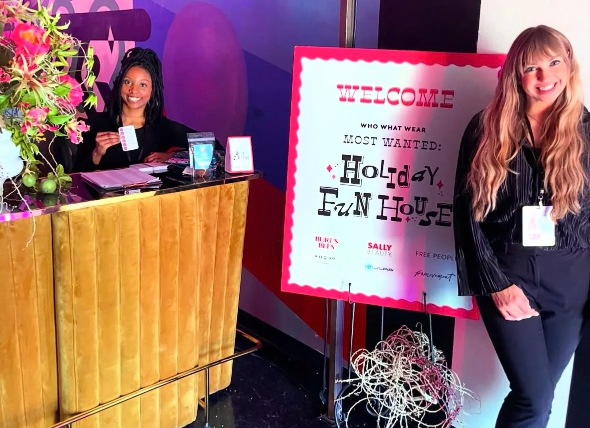 A woman at a reception desk holding a card and smiling, with a woman standing next to a colorful sign welcoming visitors to a holiday event, decorated with flowers and festive lettering.