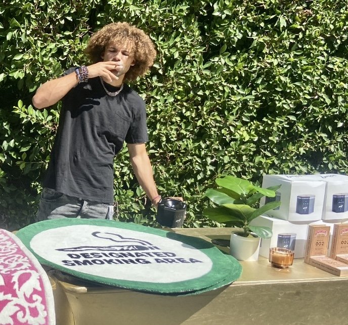 A young man with curly hair and bracelets on his wrist, standing outdoors in front of green bushes. He is holding a coffee mug in one hand and a cigarette in the other, near a table with a plant, some packaged items, and a smoker device.