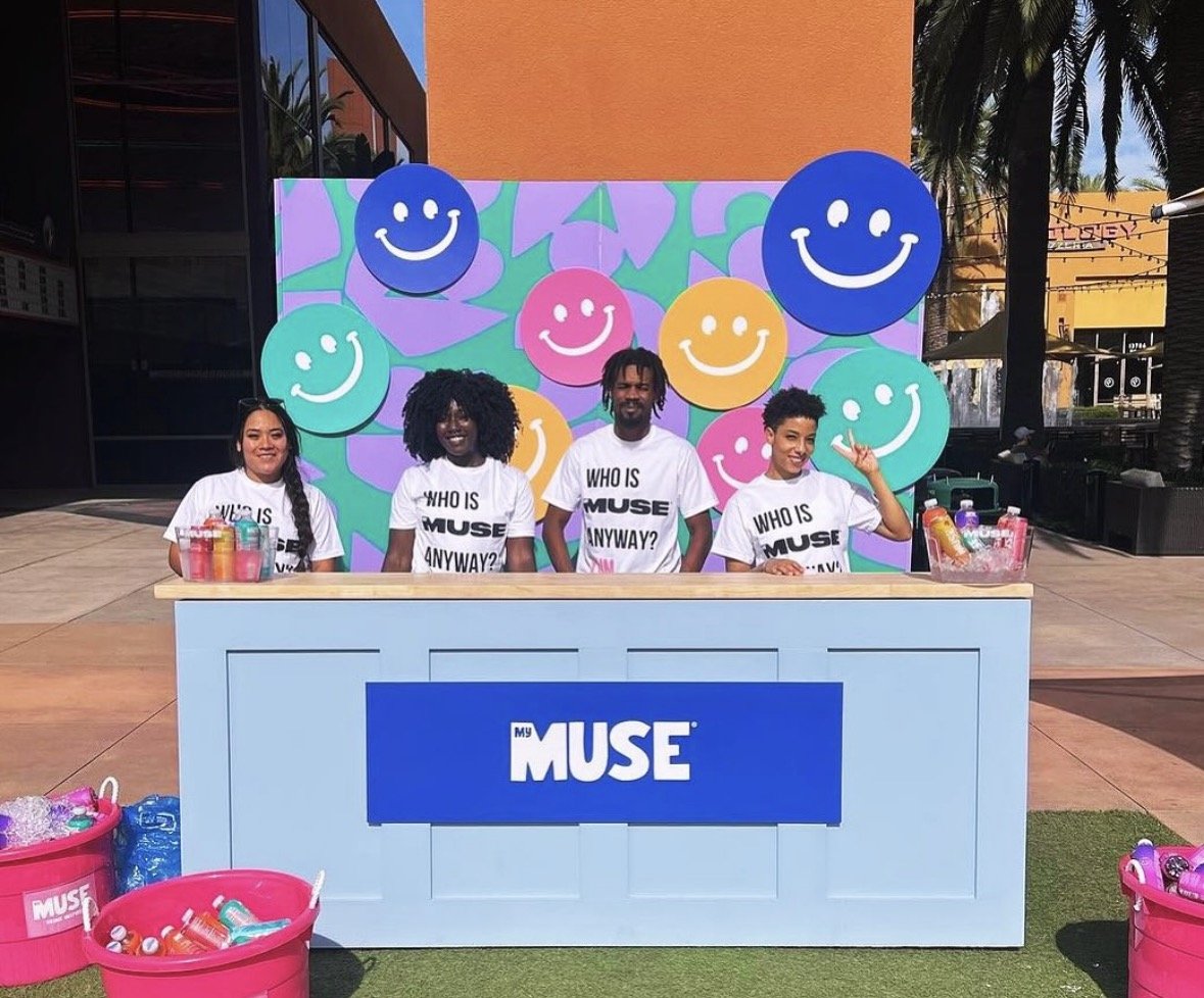 Four people standing behind a booth with a blue sign that says "My MUSE" and colorful smiley face decorations in the background. They are wearing white T-shirts with the words "Who is MUSE anyway?" and are outdoors at a sunny location.