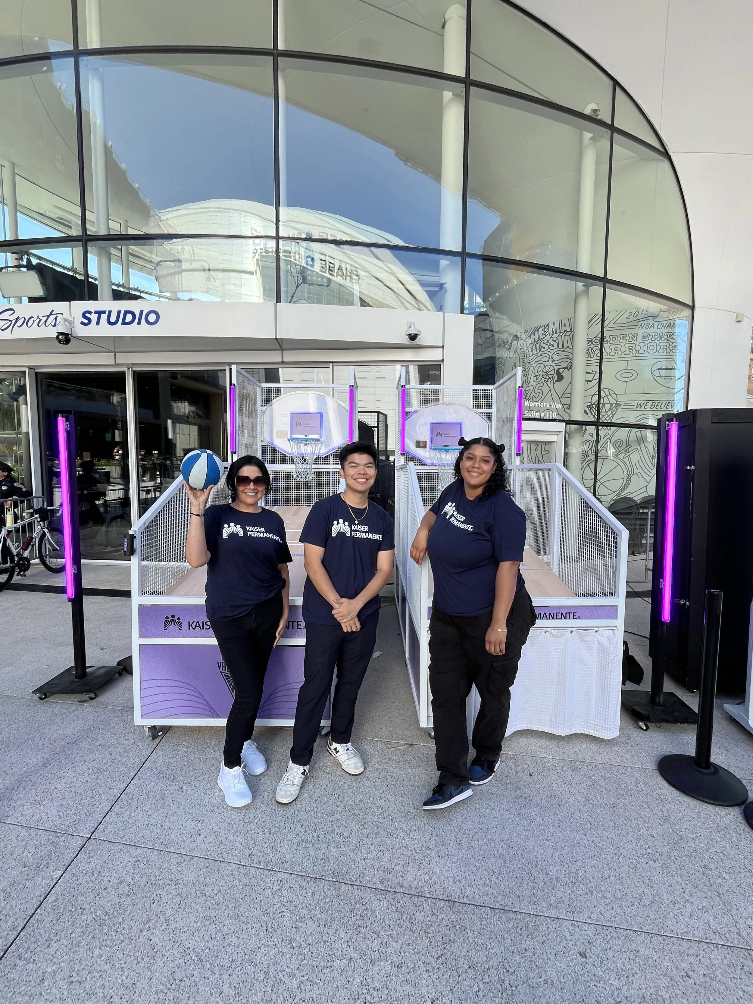 Three people wearing navy blue Kaiser Permanente shirts standing outside a glass building with purple lighting, smiling, with one holding a blue and white volleyball.