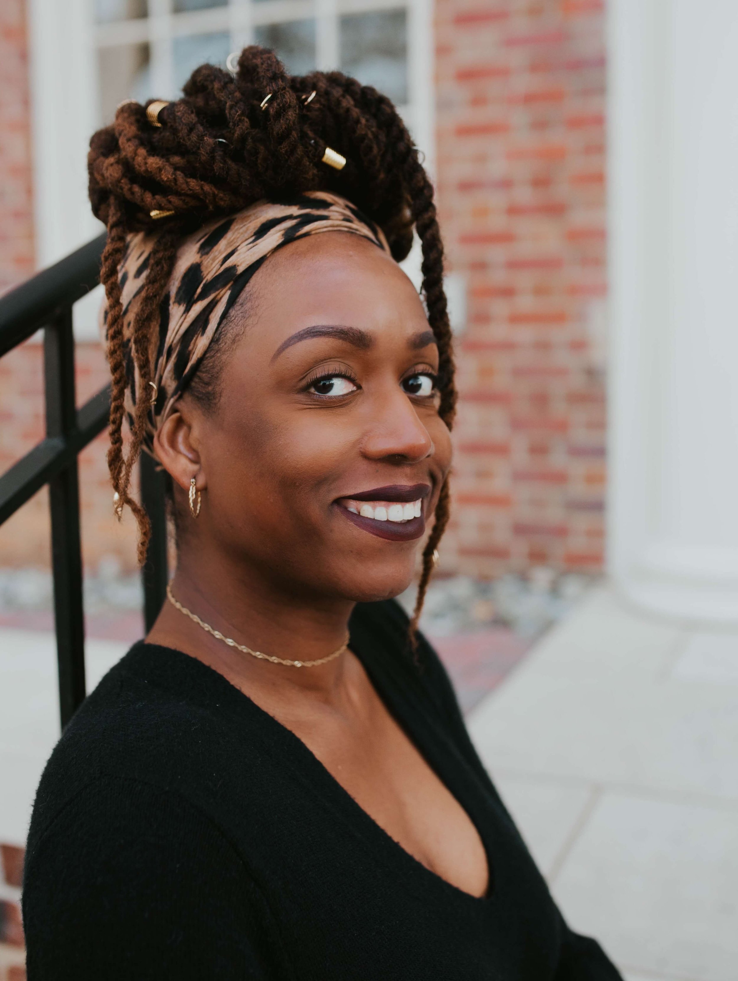Sherelle Ducksworth, wearing a leopard print headscarf, gold hoop earrings, and a gold necklace, standing outdoors in front of a brick building, smiling at the camera.