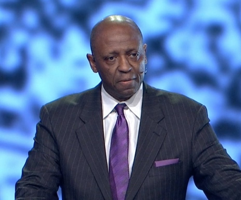 Rufus Smith in a dark pinstripe suit with a white shirt and purple tie, giving a sermon, standing in front of a blue blurred background.