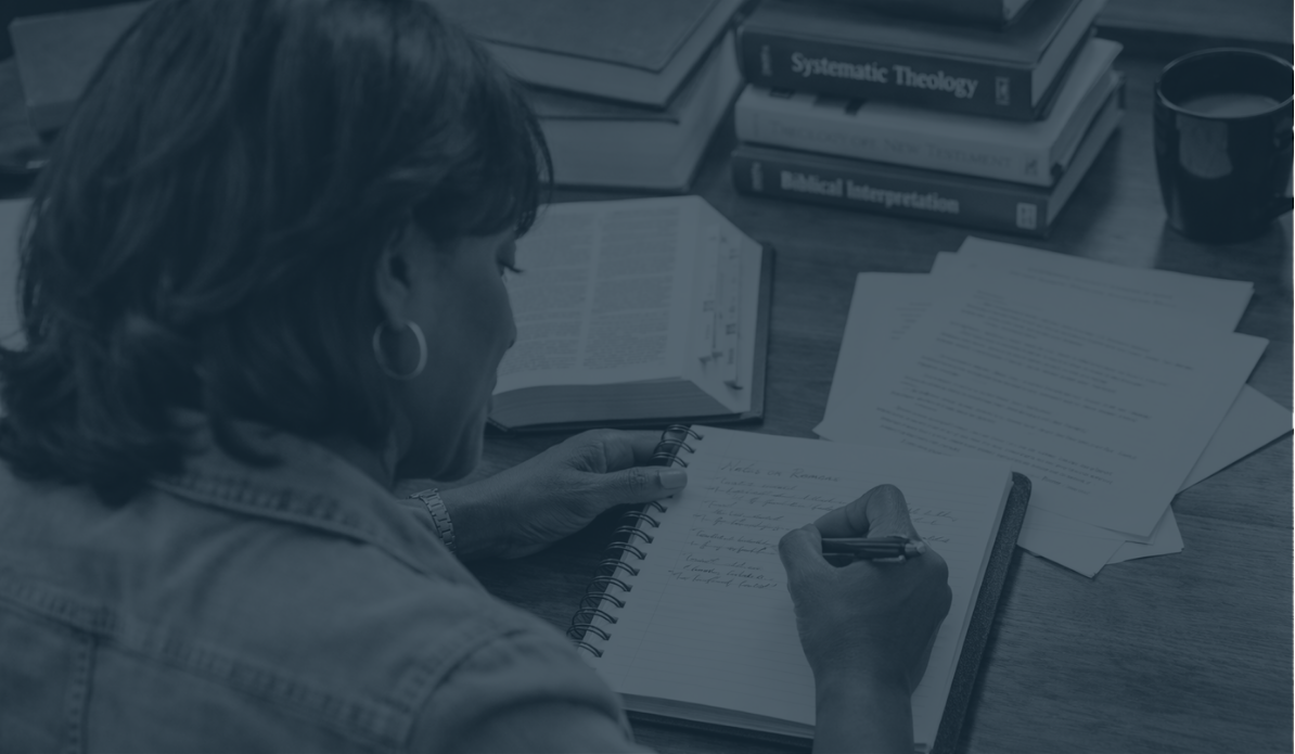 African American woman sitting at a desk writing in a notebook with a pen. There are books, papers, and a cup on the desk.