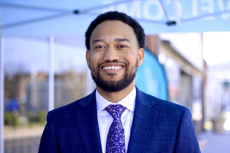 Kelvin J. Washington in a blue suit and tie standing outdoors, with a blurred background of buildings and a blue awning.