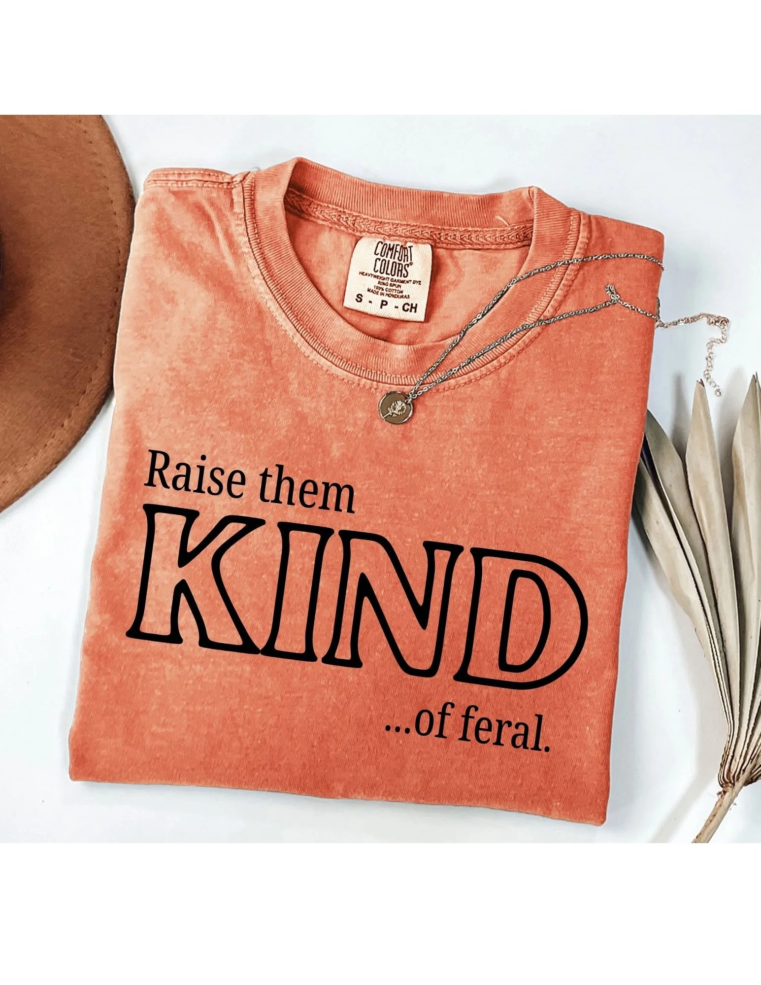 Coral-colored T-shirt with printed text and a silver necklace with a round pendant, partially visible, placed on a white surface with a brown round object and a metallic object nearby.