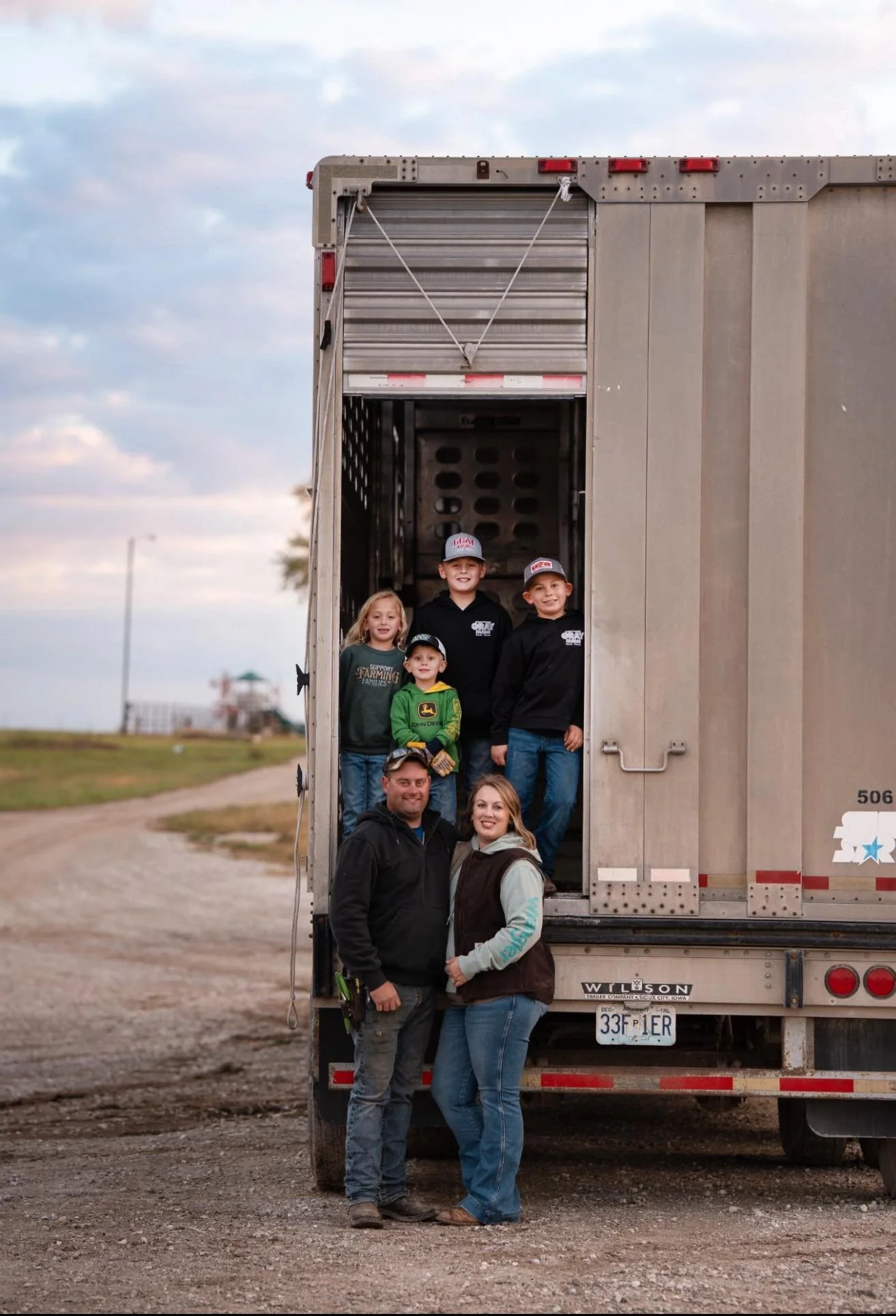 A group of seven people standing in and outside a large truck trailer, with two adults in front and five children inside the trailer. The background shows an open field with a dirt path, cloudy sky, and distant machinery.