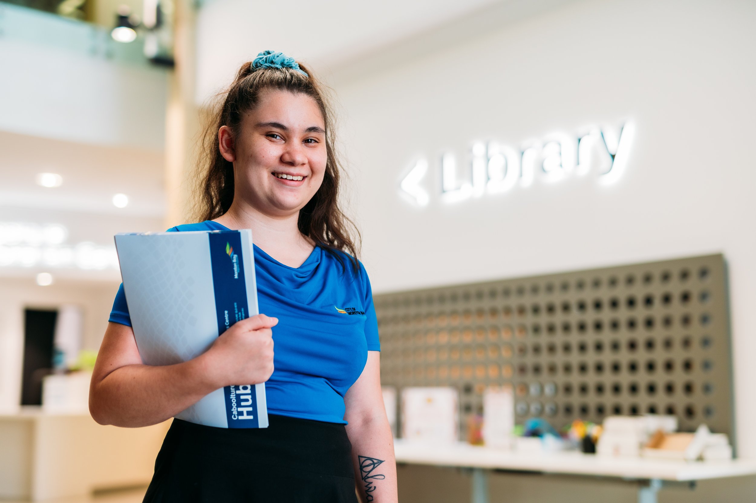 A young woman with brown hair tied with a blue scrunchie, smiling and holding a blue folder, standing in a bright, modern indoor setting.