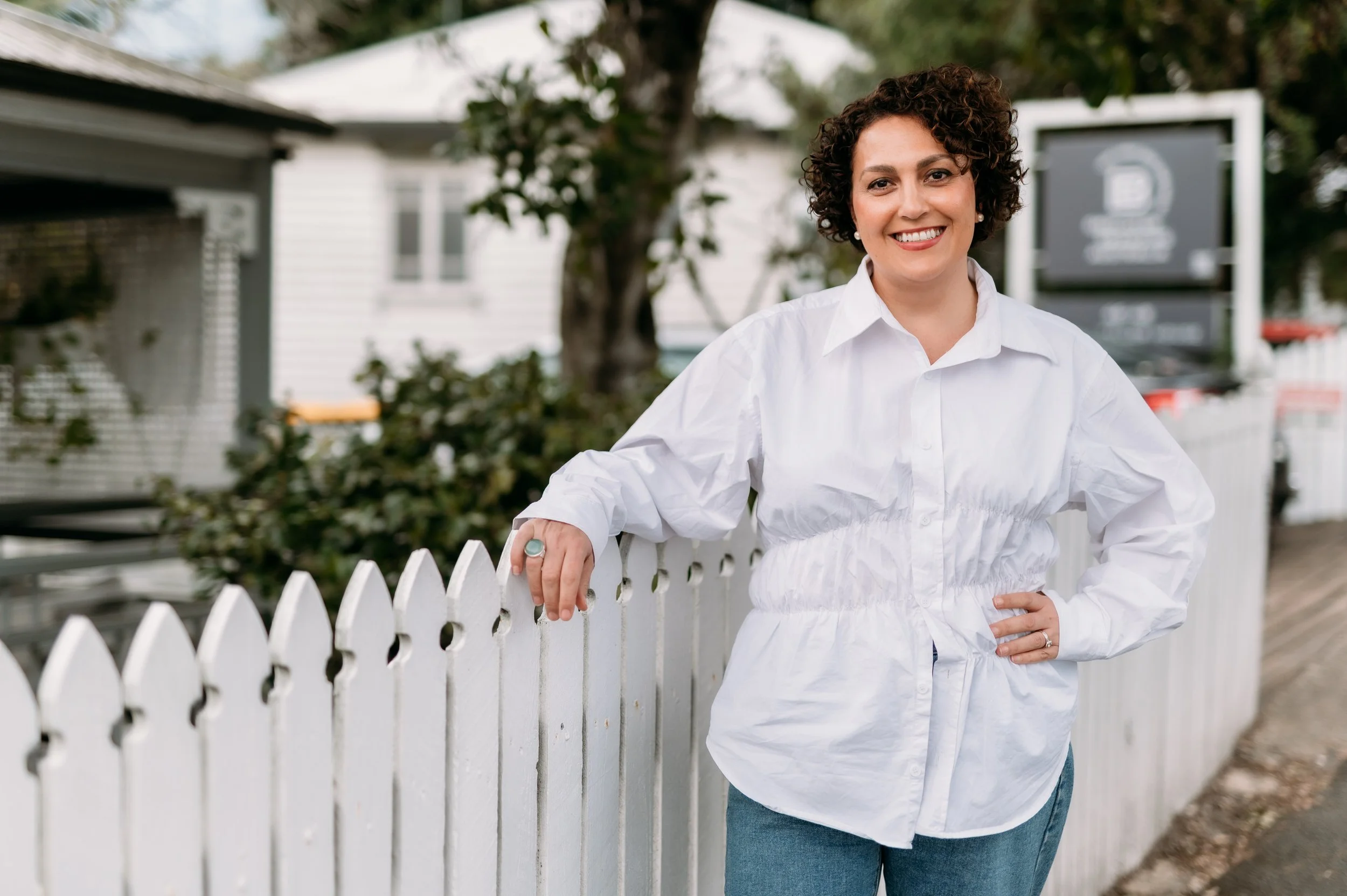 A woman with curly hair wearing a white shirt and jeans, smiling, leaning on a white picket fence outdoors.