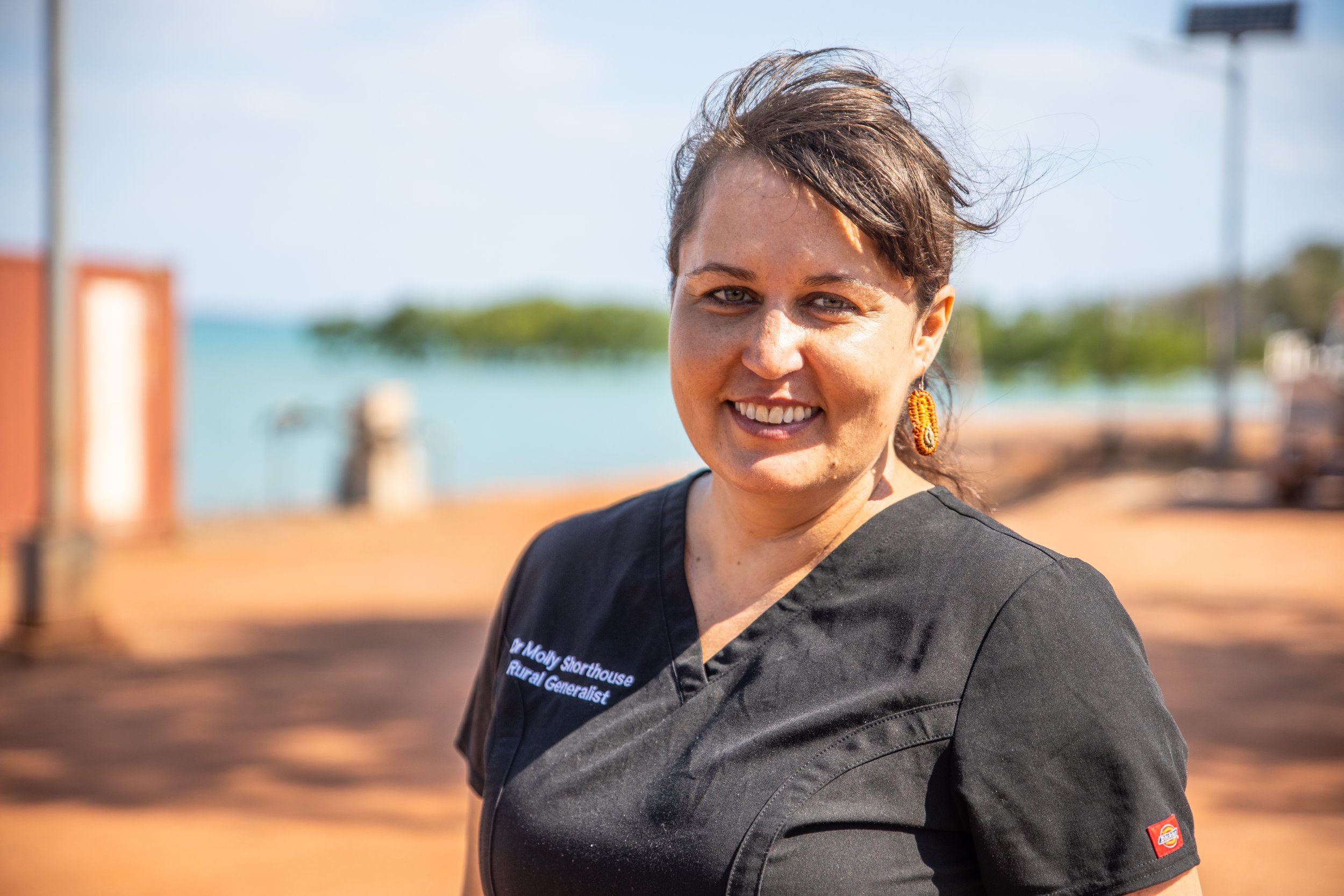 A woman smiling outdoors with short brown hair, wearing a black shirt and orange earrings, with a blurred background of a blue sky, water, and green trees.