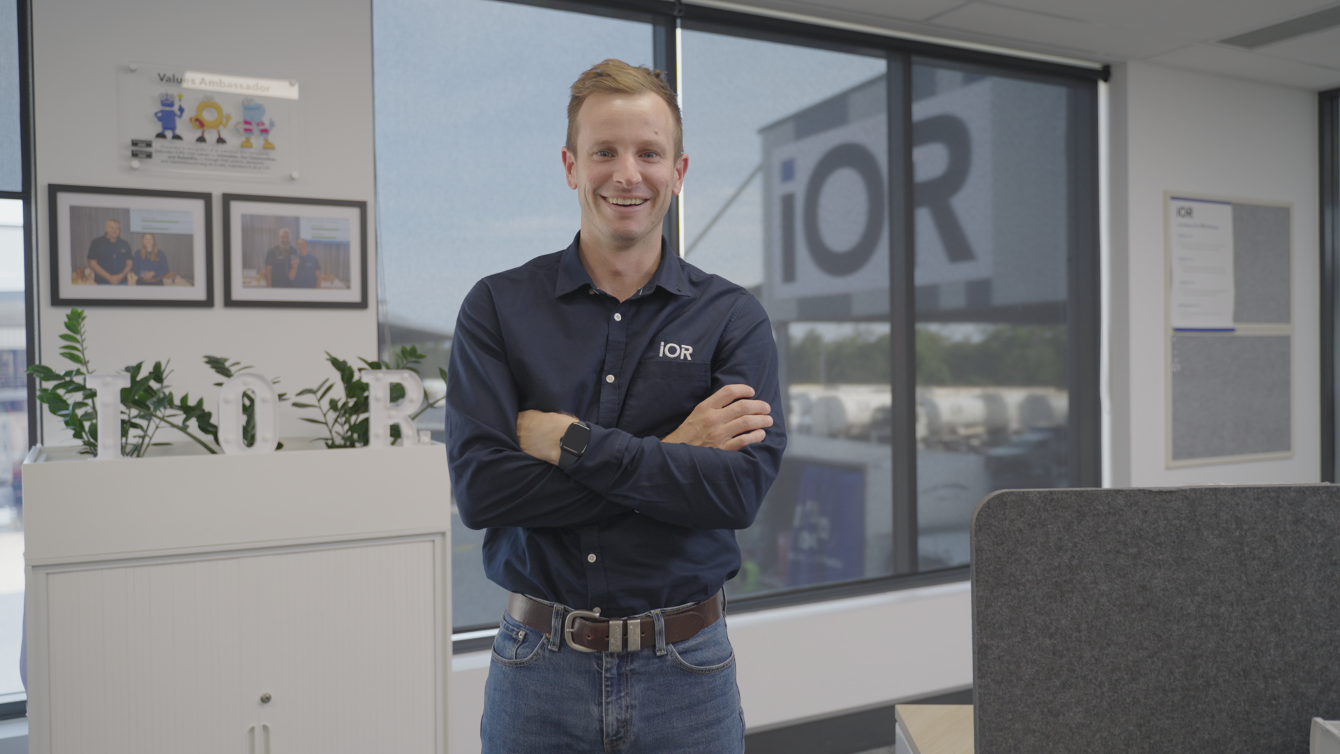 A man with light skin, short reddish hair, and wearing a dark blue shirt with 'IOR' embroidered on it, stands with arms crossed inside an office near large windows, smiling at the camera.