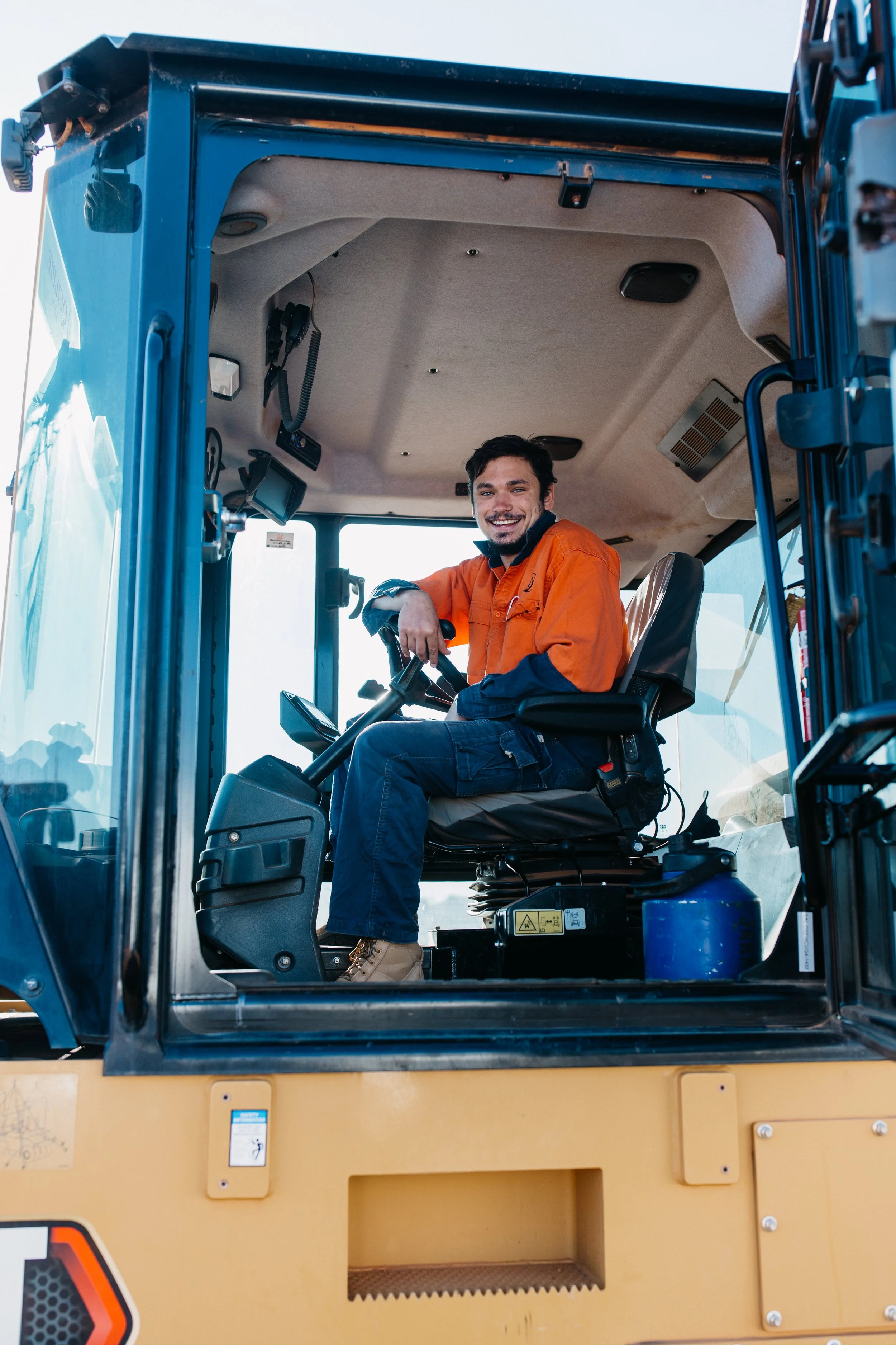 A man smiling while sitting in the driver’s seat of a construction vehicle, wearing an orange safety jacket.
